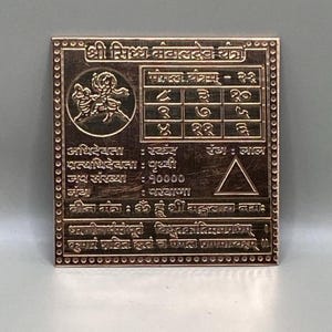 May include: A square, copper-colored metal plaque with etched text and symbols. The plaque features a grid of numbers, a triangle, and a circular image of two figures. The text is in a non-English script.