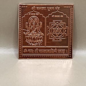 May include: A square, copper-colored plaque with intricate engravings. The design includes two main sections with detailed figures and geometric patterns, along with text in a script. The edges of the plaque have a decorative border.