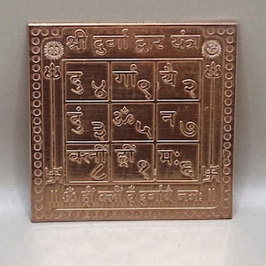 Puede incluir: Un Durga Yantra cuadrado de color cobre con intrincados diseños y texto en relieve. La cuadrícula central contiene caracteres sánscritos dentro de cuadrados individuales. El borde presenta patrones decorativos y texto adicional.