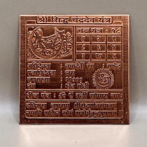 Puede incluir: Placa metálica cuadrada de color cobre con texto y símbolos grabados. La placa presenta una cuadrícula central de números, junto con una escritura detallada en un idioma que no es inglés. El diseño sugiere un artefacto espiritual o religioso.