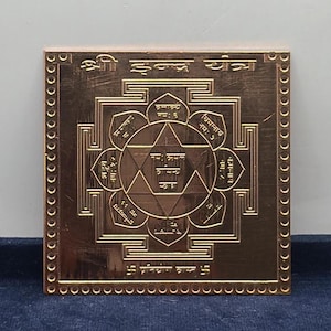 Puede incluir: Una placa metálica cuadrada de color cobre con un diseño geométrico grabado de Shri Yantra. La superficie muestra intrincados círculos concéntricos, pétalos de loto y un patrón de estrella central con texto en escritura devanagari grabado.
