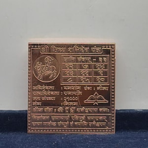 Puede incluir: Una placa metálica cuadrada de color cobre con textos en sánscrito y cuadrículas numéricas grabadas. El diseño incluye una ilustración circular de una figura montando un animal, un símbolo de arco y flecha y patrones de borde intrincados.