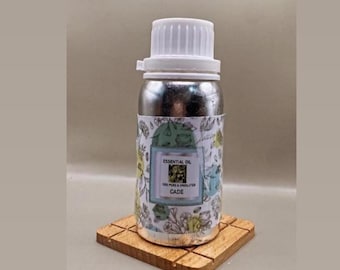 Könnte beinhalten: Eine silberne Metallflasche mit weißem Schraubverschluss und einem Etikett mit botanischem Muster, das den Text trägt: ESSENTIAL OIL, 100% PURE & UNDILUTED, CADE. Die Flasche ist zylindrisch mit einer glatten, reflektierenden Oberfläche und einem Etikett.