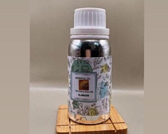 Könnte beinhalten: Eine silberfarbene Flasche mit weißem Schraubverschluss und einem Etikett mit botanischen Illustrationen sowie dem Text ESSENTIAL OIL 100% PURE & UNDILUTED AJWAIN. Die Flasche steht auf einem quadratischen Holzsockel mit eingraviertem Gittermuster.