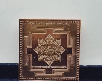 May include: A square copper-colored metal plate featuring an engraved Narsingh Pujan Yantra. The design includes a central star within a lotus flower, surrounded by geometric patterns and Sanskrit text, with a dotted border along the outer edge.