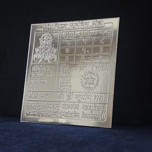 May include: A square silver-colored metal plate featuring intricate engravings of Hindu iconography, a numerical grid, and Sanskrit text. The design includes a central deity figure, a sun symbol, and various inscriptions etched into the reflective surface.
