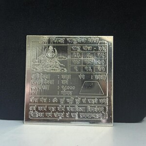 May include: A silver square with engraved text and a detailed illustration of a deity. The text includes numerical tables and Sanskrit script. The item is likely a religious or spiritual artifact.