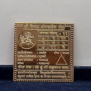 Puede incluir: Una placa metálica cuadrada de color dorado con escritura devanagari grabada, una cuadrícula numérica y una ilustración circular de una figura montando un animal. El borde está decorado con pequeñas hendiduras circulares.