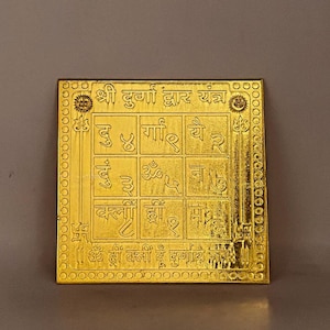 Puede incluir: Placa cuadrada dorada con intrincados patrones en relieve y texto. La placa presenta una cuadrícula de nueve cuadrados llenos de símbolos y texto en escritura. El borde está decorado con un patrón circular repetitivo. El texto en la parte superior dice "Shri Durga Dwar Yantra."