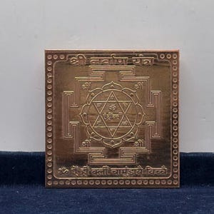 May include: A square copper-colored metal plate featuring an engraved Shri Navarna Yantra design. The center displays a six-pointed star with Sanskrit characters, surrounded by geometric patterns, circular borders, and additional Sanskrit text.