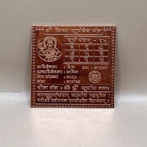 Puede incluir: Una placa cuadrada de color cobre con intrincados grabados y texto en escritura extranjera. La placa presenta una cuadrícula central, una imagen circular de una deidad y bordes decorativos. El texto incluye las palabras "सूर्य मंत्रा" y "ॐ ह्रीं सूर्याय नमः".