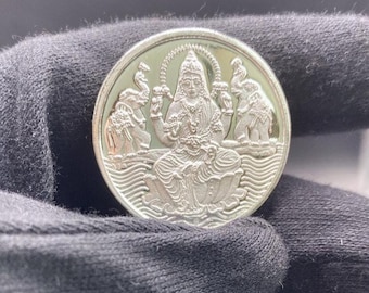 May include: A silver coin featuring a seated deity, possibly Lakshmi, adorned with intricate details. Two elephants flank the central figure, and the coin has a detailed border. The coin is held in a black glove.
