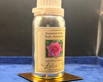 May include: A clear bottle with a white cap containing essential oil. The label features the text Essential Oil By Vedic Rudraksha, 100% Pure & Undiluted, Lotus Oil, and a graphic of a pink lotus flower on a cream background.