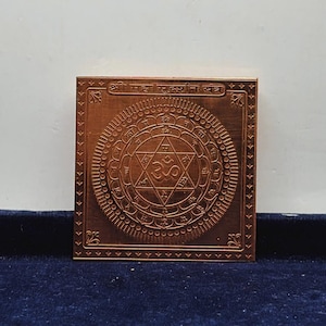 May include: A square copper-colored metal plate featuring an engraved geometric yantra design. The center displays a six-pointed star with an Om symbol, surrounded by concentric circles with Sanskrit characters and a border with decorative corner motifs.