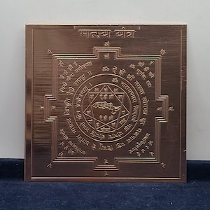 Può includere: Una piastra metallica quadrata color rame con un intricato disegno geometrico inciso. Il centro mostra un motivo a stella con un simbolo di pesce, circondato da cerchi concentrici e testo in sanscrito. Il bordo superiore include il testo Matsya Yantra.