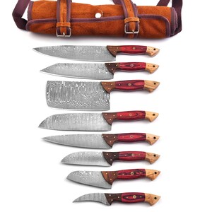 May include: A set of eight stainless steel kitchen knives with patterned blades and wooden handles with red accents. The knives are displayed with a brown leather knife roll with buckle closures.