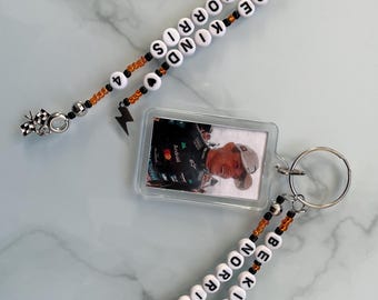 Puede incluir: Dos llaveros con colgantes rectangulares transparentes con imágenes de una persona. Cada llavero tiene una hebra de cuentas con las palabras "BE KIND NORRIS 4" deletreadas en cuentas de letras blancas. Un llavero tiene un dije de bandera a cuadros, el otro un rayo.
