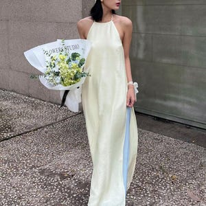 Könnte beinhalten: Eine Frau trägt ein cremefarbenes Neckholder-Kleid über hellblauen Hosen, ein gehäkeltes Stirnband und weiße Schuhe. Sie hält einen Blumenstrauß, der in weißes Papier mit dem Text "FLOWERS STUDIO" gewickelt ist. Das Kleid hat einen hohen Schlitz.