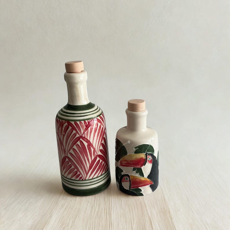 Handmade ceramic olive oil and vinegar bottles with cork stoppers, tropical toucan and red palm leaf designs on neutral background.
Artisan pottery cruet set, hand-painted Mediterranean style, unique ceramic dispensers for kitchen decor.
Decorative ceramic oil bottles, rustic boho style kitchenware, perfect for olive oil, vinegar, or balsamic.
