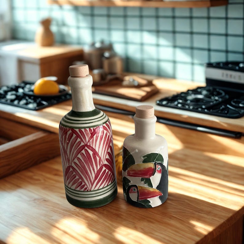 Hand-painted ceramic oil and vinegar bottles with red palm leaf and tropical toucan motifs, displayed on a wooden kitchen counter under natural light — artisan Mediterranean-style pottery perfect for boho kitchens, eco-friendly dining, or as a creative chef’s gift.