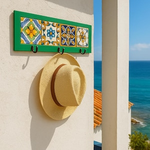 Könnte beinhalten: Ein grüner Wandhaken aus Holz mit vier dekorativen Keramikfliesen mit floralen Mustern in Gelb, Blau und Weiß. Ein Strohhut mit braunem Band hängt unter den Haken. Die Szene spielt vor dem Hintergrund des Ozeans und des blauen Himmels.