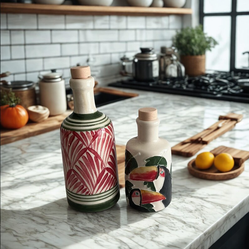 Hand-painted ceramic cruet bottles with tropical toucan and palm leaf patterns, displayed on a marble kitchen countertop with lemons and wooden accessories — vibrant Mediterranean-style pottery ideal for olive oil, vinegar, or as a stylish boho kitchen gift.