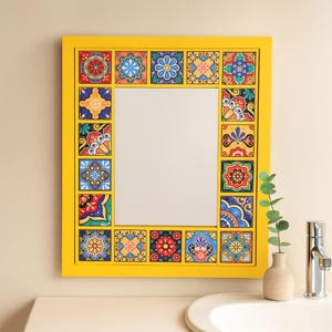 Puede incluir: Un espejo cuadrado con un marco amarillo brillante. El marco está decorado con azulejos coloridos y estampados. El espejo está colgado sobre un lavabo blanco con un grifo plateado y un pequeño jarrón con vegetación.