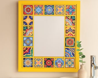 May include: A square mirror with a bright yellow frame. The frame is decorated with colourful, patterned tiles. The mirror is hanging above a white sink with a silver tap and a small vase with greenery.