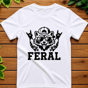 May include: White t-shirt featuring a black graphic of a raccoon wearing sunglasses and a backwards baseball cap, with the word "FERAL" in bold letters below. The raccoon is making a rock and roll hand gesture.