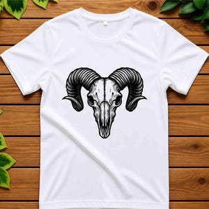 May include: White t-shirt featuring a detailed black and white illustration of a ram skull. The skull has large, curved horns and intricate shading, creating a striking contrast against the white fabric. The design is centered on the shirt.