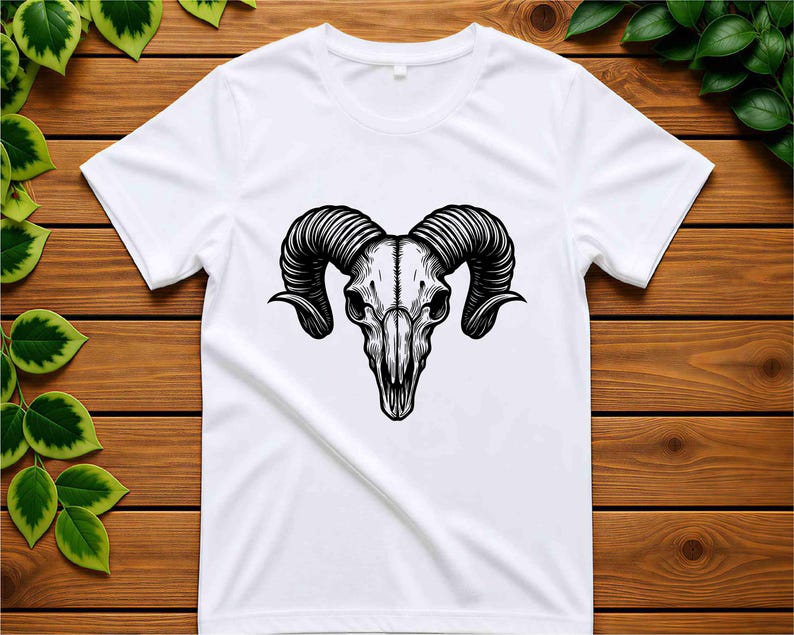 May include: White t-shirt featuring a detailed black and white illustration of a ram skull. The skull has large, curved horns and intricate shading, creating a striking contrast against the white fabric. The design is centered on the shirt.