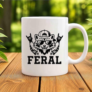 May include: White ceramic mug featuring a black graphic of a raccoon wearing sunglasses and a backwards baseball cap, with the word "FERAL" in bold letters below. The raccoon is making a rock and roll hand gesture.