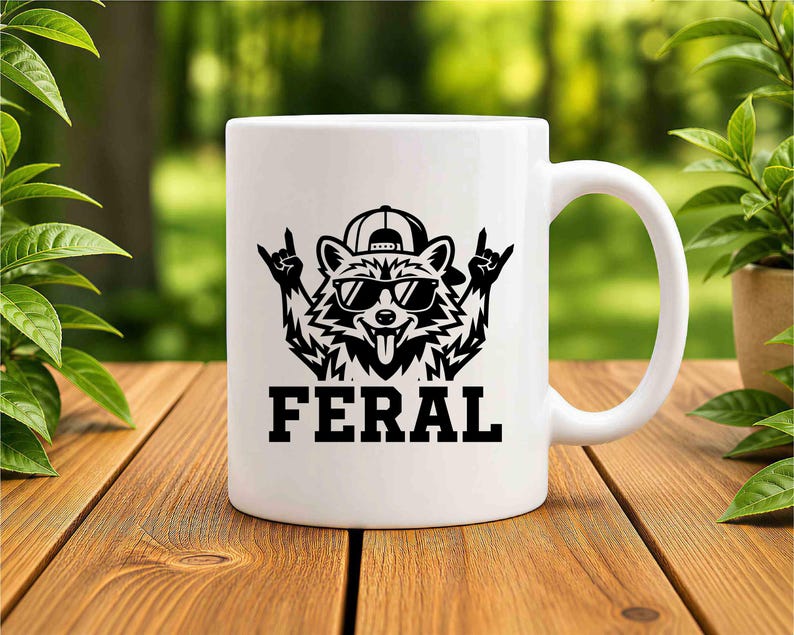 May include: White ceramic mug featuring a black graphic of a raccoon wearing sunglasses and a backwards baseball cap, with the word "FERAL" in bold letters below. The raccoon is making a rock and roll hand gesture.