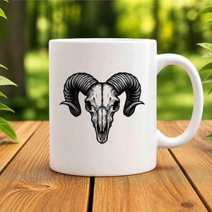 May include: White ceramic mug featuring a detailed black illustration of a ram skull. The skull has large, curved horns and is centered on the mug. The mug has a standard handle and is set on a wooden surface.