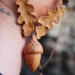 May include: A handcrafted wooden necklace featuring an acorn pendant and two oak leaf charms. The pendant and charms are carved from light-colored wood and suspended from a black cord with wooden beads. The necklace is a nature-inspired accessory.