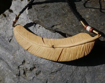Op de afbeelding: Een houten veer hanger ketting. De hanger is lichtbruin van kleur met gedetailleerde gesneden lijnen, die op een veer lijken. De ketting heeft een zwart gevlochten koord met messing en witte accenten.