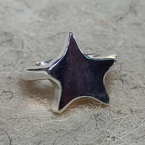Puede incluir: Un anillo plateado brillante con forma de estrella. El anillo presenta un diseño de estrella de cinco puntas con una superficie lisa y pulida. El anillo está sobre un fondo texturizado de color claro.