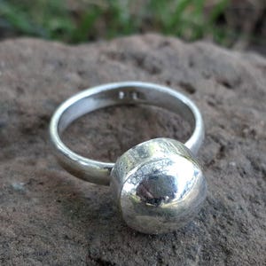 May include: A silver ring with a large, round, polished stone. The ring band is smooth and simple, leading to the prominent, spherical stone. The ring is set against a textured, gray-brown stone surface.