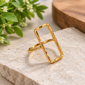 Puede incluir: Un anillo de tono dorado que presenta un marco rectangular grande y abierto con una superficie martillada y texturizada. El anillo tiene una banda delgada y lisa que se conecta a la base del rectángulo vertical, creando un diseño geométrico audaz.
