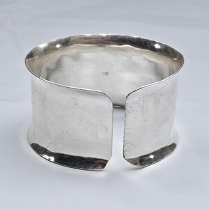 Puede incluir: Una pulsera de brazalete abierta y ancha hecha de plata pulida con una textura de superficie lisa y ligeramente irregular. El interior de la banda presenta una pequeña marca numérica estampada 925, que indica el contenido de plata del metal.