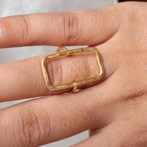 Pode incluir: Um anel dourado com um design retangular. A forma do anel é um retângulo oco com cantos arredondados. O anel é usado em um dedo, mostrando sua estética simples, mas moderna. O anel é feito de metal.