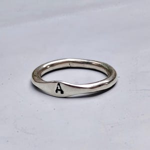 Puede incluir: Un anillo de metal plateado con una banda lisa y redondeada y una sección frontal ligeramente aplanada. La letra A está grabada en negro en el centro de la parte aplanada del anillo, que tiene un acabado de superficie pulido y brillante.
