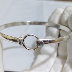 May include: A silver bangle bracelet with a circular clasp. The bracelet is round and has a simple, elegant design. The clasp is a small circle that connects to the other side of the bracelet. The bracelet is held by a person wearing white gloves.