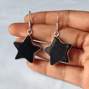 Peut inclure: Une paire de boucles d'oreilles pendantes avec des pierres noires en forme d'étoile serties dans des montures en métal argenté. Chaque boucle d'oreille est suspendue à un crochet simple, présentant un design d'étoile à cinq branches.