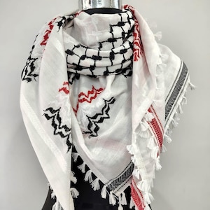 Puede incluir: Bufanda blanca con patrones geométricos negros y rojos y borlas decorativas. La bufanda está colocada sobre un maniquí, mostrando su diseño y textura. La bufanda tiene un patrón de cuadros y diseños en zigzag.