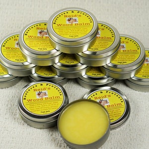 May include: A stack of small, silver tins containing Reid's Wood Balm. The tins are labeled with yellow and black text that reads "Restores & Protects Reid's Wood Balm". The balm is food safe and is used to restore and protect wood.