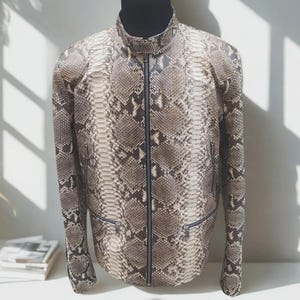 May include: A snakeskin-effect jacket with a stand-up collar and a full-length zip. The jacket features two zipped pockets and is displayed on a black mannequin. The colour palette includes shades of brown, tan, and grey.