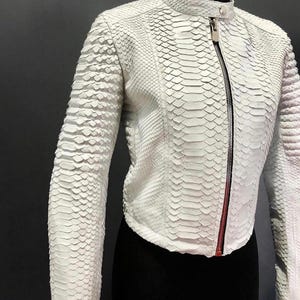 May include: A white leather jacket with a snakeskin texture. It has a stand-up collar, long sleeves, and a full-length zip with a red accent. The jacket is displayed on a black mannequin.