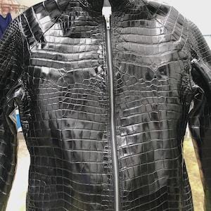 Pode incluir: Casaco de couro preto com um padrão texturizado semelhante à pele de crocodilo. O casaco tem gola alta, zíper completo e mangas compridas. O acabamento brilhante realça a textura detalhada do material.