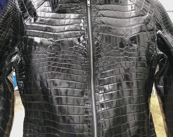 Può includere: Giacca in pelle nera con texture a pelle di coccodrillo. La giacca ha un collo alto, una cerniera a tutta lunghezza e una silhouette aderente. La pelle ha una finitura lucida e la giacca sembra nuova.